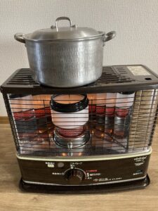 石油ストーブの上に置かれた、蓋の閉まった銀色の鍋。冬の室内で牛筋大根をコトコト煮込んでいる様子。