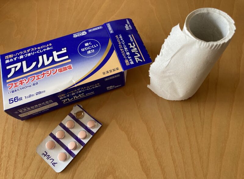 花粉症の市販薬「アレルビ」と使いかけのトイレットペーパーが置かれた机の様子。重症者のリアルな日常を表現。