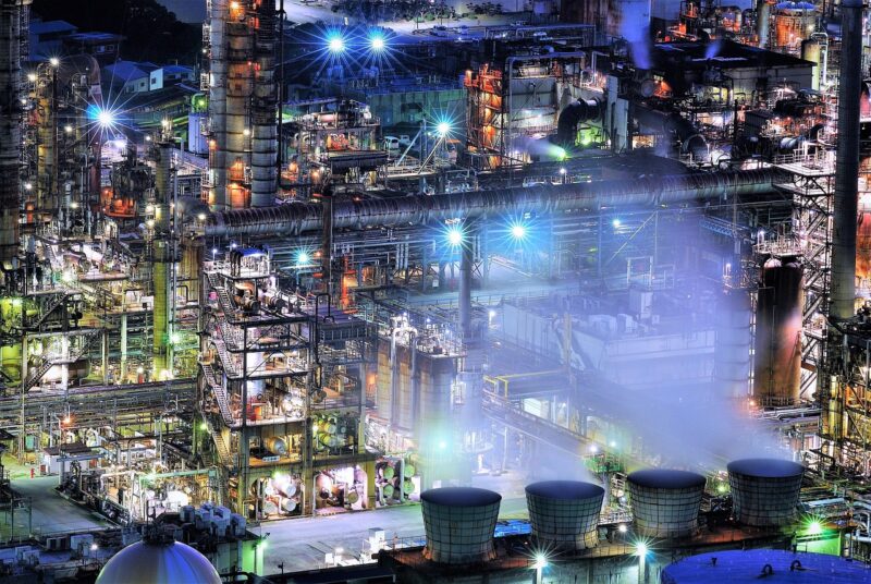 夜間に煌々と明かりが灯る大規模な工場の夜景。複雑に入り組んだ配管や巨大な煙突が、暗闇の中でライトアップされている。工場勤務の夜勤に関する記事の文中で、その独特な風景を伝えるために使われている画像。