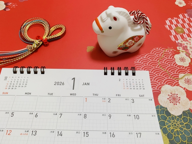 赤い背景に置かれた2026年1月の卓上カレンダーとお正月の縁起物。花粉症の早期対策が必要な時期を視覚的に表現している。