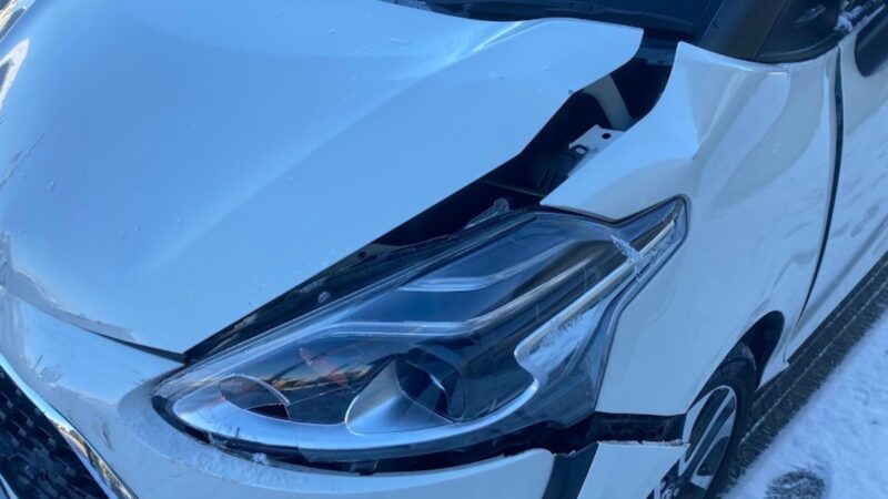 雪が残る路面でスリップし、ゴミ収集車と衝突した白いトヨタ・シエンタのフロント部分の近接写真。ヘッドライトが割れ、ボンネットが大きく歪んだ事故直後の生々しい損傷の様子。