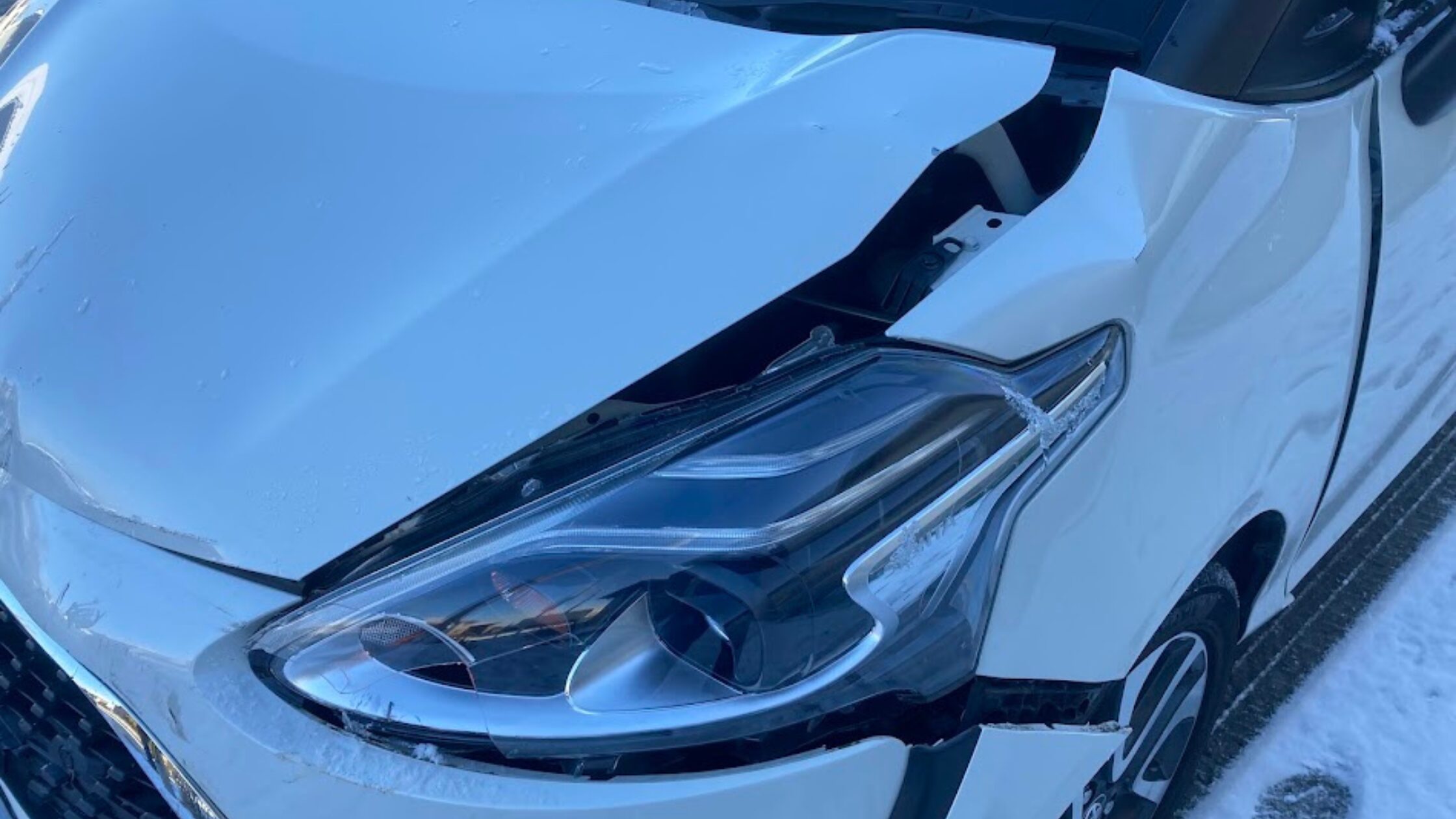 雪が残る路面でスリップし、ゴミ収集車と衝突した白いトヨタ・シエンタのフロント部分の近接写真。ヘッドライトが割れ、ボンネットが大きく歪んだ事故直後の生々しい損傷の様子。