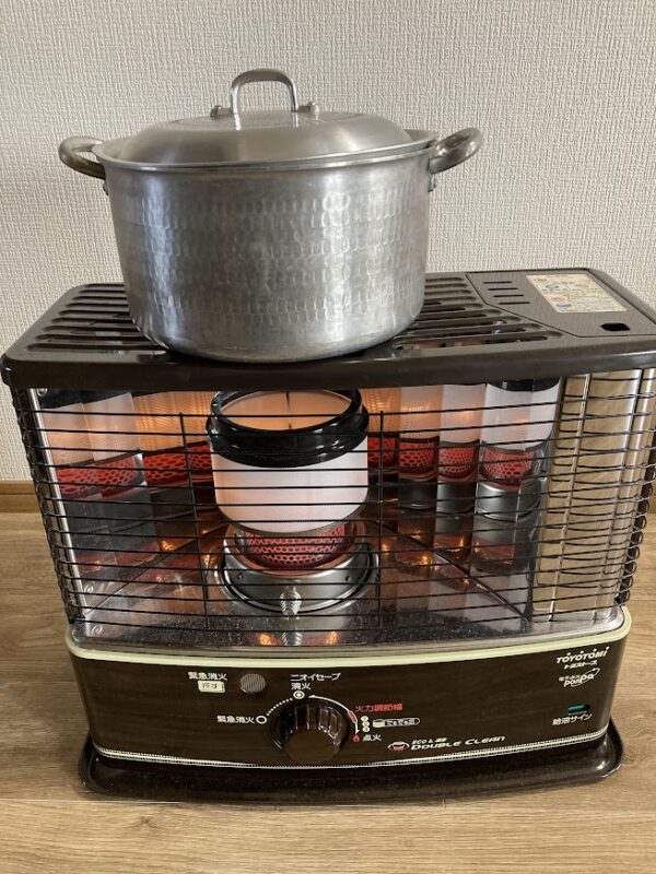 石油ストーブの上に置かれた、蓋の閉まった銀色の鍋。冬の室内で牛筋大根をコトコト煮込んでいる様子。