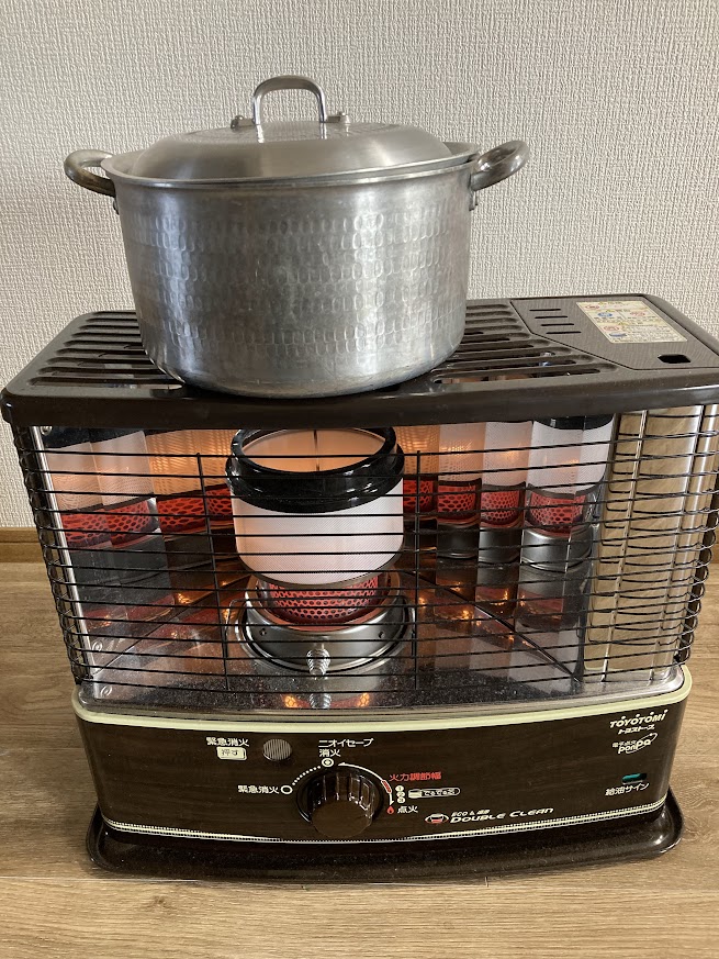 石油ストーブの上に置かれた、蓋の閉まった銀色の鍋。冬の室内で牛筋大根をコトコト煮込んでいる様子。