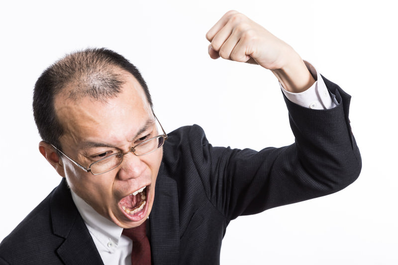 眼鏡をかけたスーツ姿の男性が、拳を握りしめ、顔を真っ赤にして激しい形相で怒鳴り散らしている様子