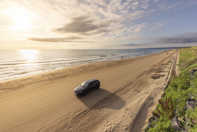 夕暮れの石川県・千里浜なぎさドライブウェイ（日本唯一の車で走れる砂浜）を走る1台の黒いSUV。波打ち際と広大な砂浜、沈みゆく夕日の美しい風景。ドローン撮影風の上空からの写真。