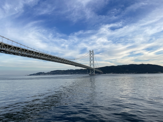 阪九フェリーの船上から見上げる、巨大な明石海峡大橋。青空に広がる白い雲と、穏やかな海面に映る橋の影。北陸から九州への旅路で撮影した、情緒あふれる風景。
