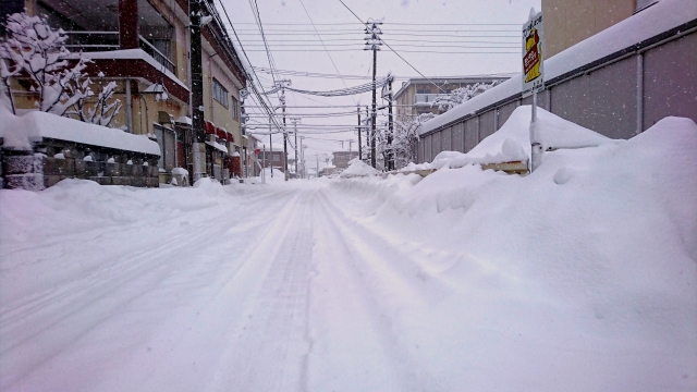 北陸地方の豪雪地域の住宅街。道路の両脇に高く積もった雪の壁と、深々と降り積もる新雪。鉛色の空と電線が広がる、雪国の日常風景。車視点からの写真。