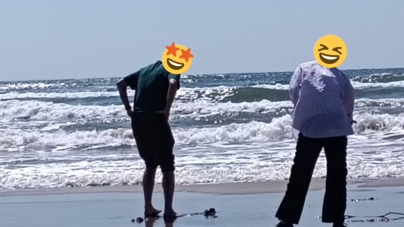 晴天の千里浜なぎさドライブウェイの波打ち際で、笑顔のスタンプで顔を隠した夫婦が裸足になって海を眺めている後ろ姿