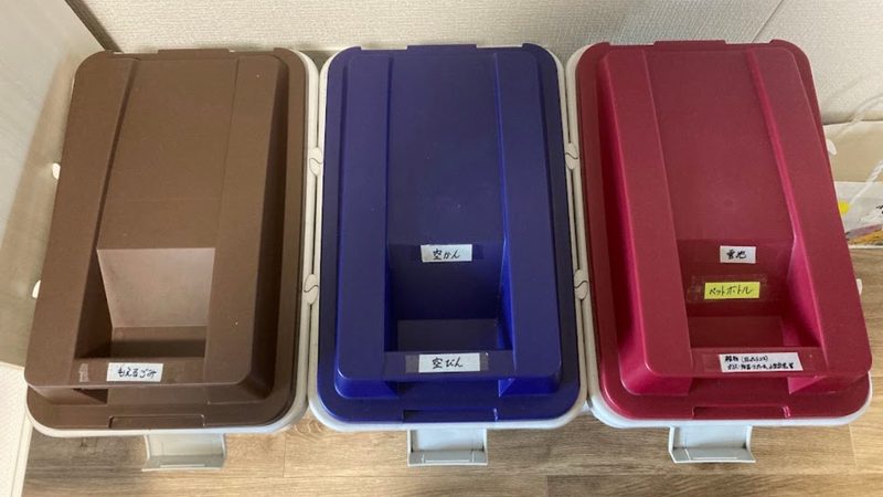 真上から見た3色の連結ゴミ箱。茶色がもえるごみ、青が空かん・空びん、赤がペットボトル・電池・雑物と分別ラベルが貼られている