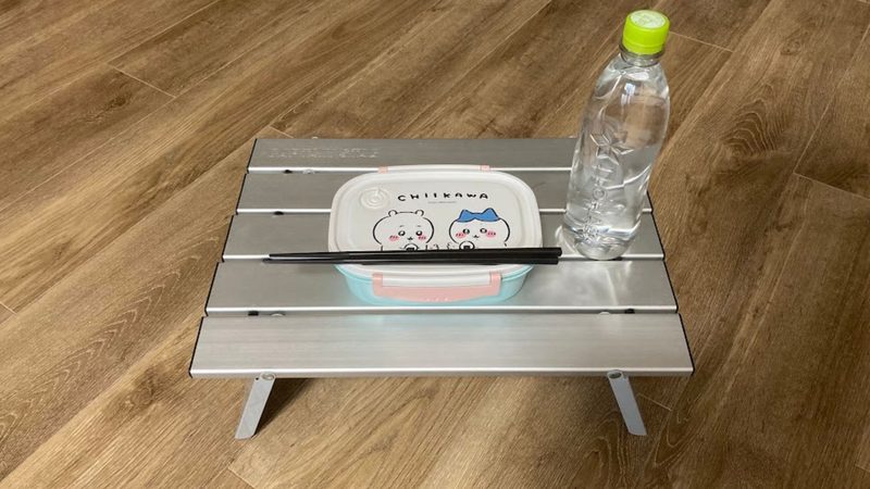 キャプテンスタッグのアルミロールテーブルのサイズ感。フローリングの床の上に置かれたミニテーブルの上に、ちいかわのお弁当箱とお箸、ペットボトルが置かれている様子。
