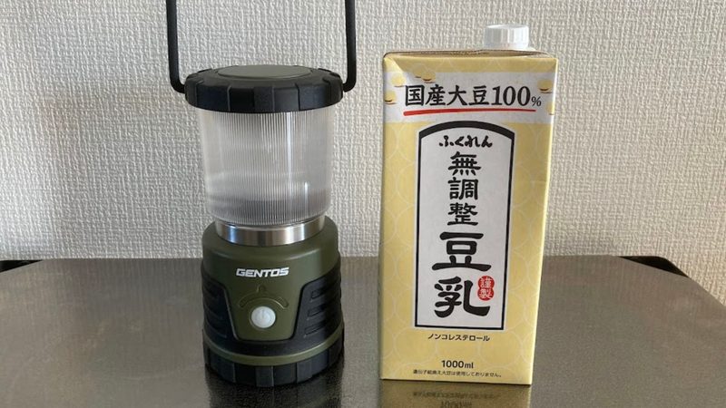 テーブルの上に置かれた点灯していないGENTOS(ジェントス)電池式ランタン(オリーブグリーン)と、その横に並べて立てられた1000mlのふくれん無調整豆乳(1Lパック)のサイズ比較写真。