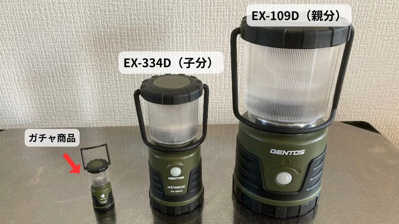 GENTOS(ジェントス)LEDランタンのサイズ比較画像。右から大型のEX-109D(ラベル:親分)、中型のEX-334D(ラベル:子分)、そして左端に赤矢印で指された超小型のガチャ商品(ミニチュア)が順に並べられている様子。