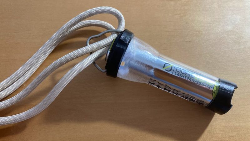 木目のテーブルの上に置かれた Goal Zero(ゴールゼロ)LIGHTHOUSE micro FLASH の金属フック部分に、首や肩から下げられるアイボリー色のパラコードが取り付けられている状態の写真。