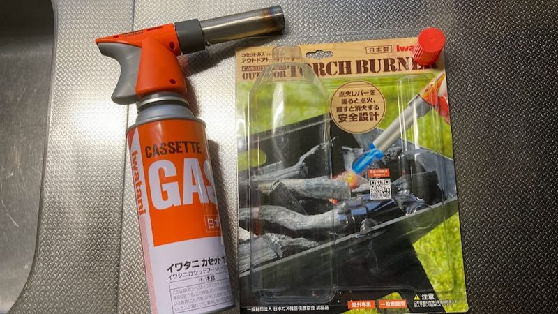 ステンレスのキッチンシンク横に置かれたイワタニ(Iwatani)のカセットガストーチバーナー。オレンジと灰色のバーナー本体がカセットボンベに装着された状態と、その隣に「点火レバーを握ると点火、離すと消火する安全設計」と書かれた台紙パッケージが並んでいる様子。