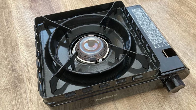 木目の床の上に置かれたIwatani(イワタニ)のカセットコンロ「カセットフータフまる(黒)」の本体写真。頑丈な黒い五徳とダブル風防ユニット、火口の焼き色が見える斜め上からのアングル。