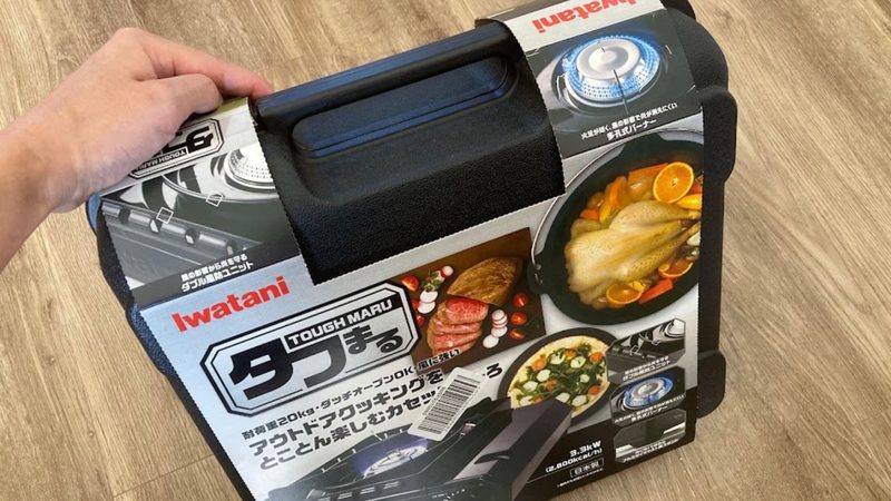 Iwatani(イワタニ)のカセットコンロ「タフまる」の専用ハードケース。紙のパッケージ帯が巻かれた状態のケースを、大人が片手で持ち上げて持ち運びやすさを示している様子。